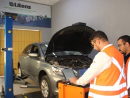 alt=homens com coletes laranjas dentro de uma oficina mecânica utilizam o scanner em um carro