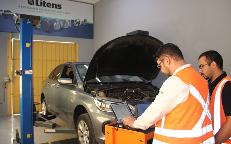 alt=homens com coletes laranjas dentro de uma oficina mecânica utilizam o scanner em um carro