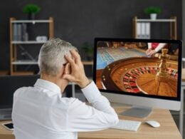 alt= homem sentado e de costas coloca as mãos na cabeça ao olhar para uma roleta de cassino na tela do computador