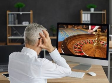 alt= homem sentado e de costas coloca as mãos na cabeça ao olhar para uma roleta de cassino na tela do computador