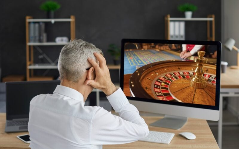 alt= homem sentado e de costas coloca as mãos na cabeça ao olhar para uma roleta de cassino na tela do computador