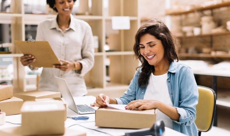 Happy young businesswoman sticking a barcode label on a package box. Online store owners preparing an order for shipping in a warehouse. Female