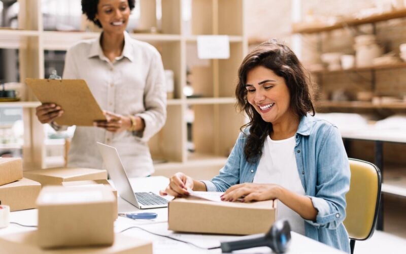 Happy young businesswoman sticking a barcode label on a package box. Online store owners preparing an order for shipping in a warehouse. Female